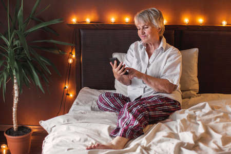 Smiling mature woman holding phone, using mobile device apps, looking at screen, happy older female chatting online, texting, writing message on cellphone, having fun at home, sitting on bedの写真素材
