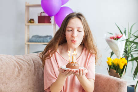 Cheerful gorgeous young woman blowing out the candles on the birthday cake. Happy teen girl celebrating her birthday at decorated homeの写真素材