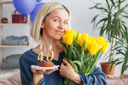 Smiling Middle aged woman with birthday cake and bouquet of flowers. Adult female celebration her birthday at homeの写真素材