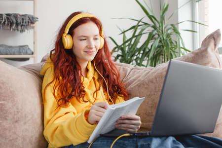 Teen girl learning language online, using laptop, looking at screen, student doing school tasks at home, writing notes, young woman listening to lecture at home, distance educationの写真素材