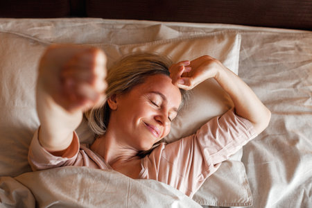 Good morning, new day, weekend, holiday. Happy middle aged woman lying on bed, lady stretching arms after sleep and enjoying morning in cozy comfort bedroom interior, free spaceの写真素材