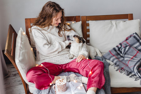 Young woman and her adorable jack russell terrier dog sitting on couch cozied up, covered with blanket. Lazy afternoon at home with loved pet concept. Close up, copy space, interior backgroundの写真素材