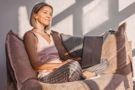 Charming middle-aged woman spends leisure time in networks on the computer, texting messages, watching movies. The adult lady works remotely with a laptop sitting on the sofa in cozy living roomの写真素材