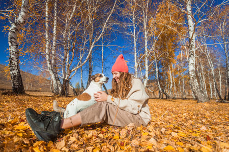 Young cheerful girl with dog walks in autumn park. Teenage girl in orange hat plays with yellow leaves in the forestの写真素材