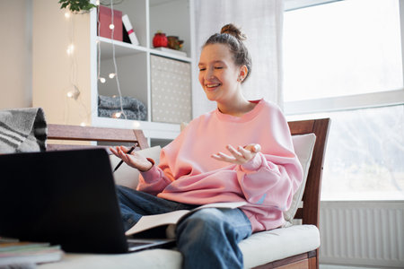 Teenager girl in an online lesson in high school. Teen is sitting on the couch with laptop, listening to the teacher, writing in a notebook, student answers a teacher's question in an online lessonの写真素材