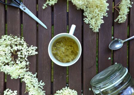 Elderberry blossom flatlay on wooden table, cup of tea, fresh herbal blossom, glass with dried elder, tea spoon, scissors.の写真素材