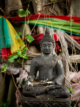 Ancient Black Buddha Statue with Colorful Prayer Flags at Sacred Banyan Tree - Buddhist Spiritual Worship Site.の写真素材