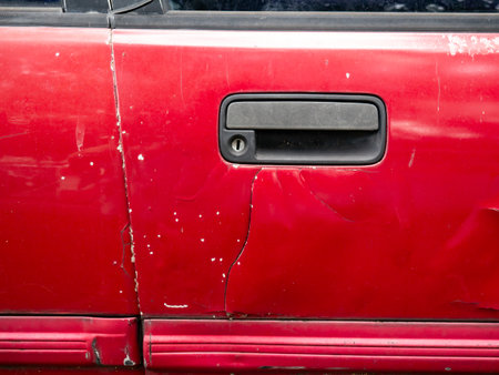 Severely damaged red car door with deep dents scratches and cracked paint near door handle from collision impactの写真素材
