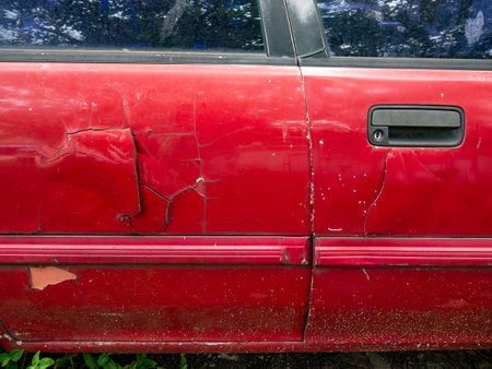 Severely damaged red car door with deep dents scratches and cracked paint near door handle from collision impactの写真素材