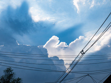Dramatic blue sky with white cumulus clouds and electric power lines crossing diagonally in urban settingの写真素材