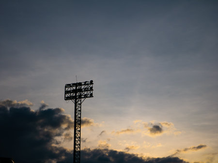Silhouette of Stadium Floodlight Tower Against Dramatic Sunset Sky with Golden Clouds at Duskの写真素材