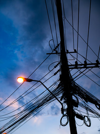 Silhouette of Electric Power Pole with Complex Cable Network Against Dramatic Blue Twilight Sky with Street Lightの写真素材