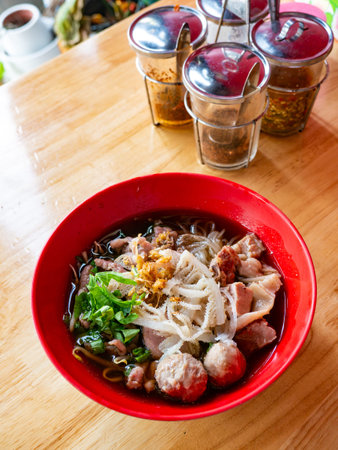 Thai Beef Noodle Soup with Meatballs Offal and Crispy Pork Skin in Red Bowl - Traditional Asian...