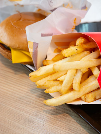 Close up crispy french fries in red paper box with cheeseburger background on tray, fast food side dish and unhealthy snack concept.