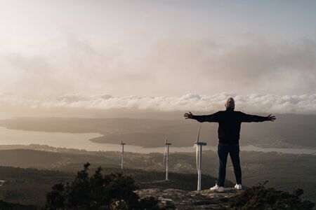 Young man stands on cliff with view of field with wind turbines at sunset in northern Spainの写真素材