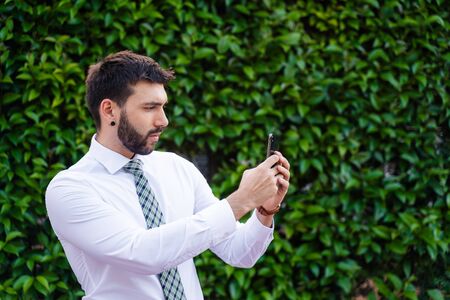 Young business man with white shirt and tie in outdoor park taking a photo with phoneの写真素材