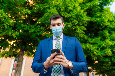 Young elegant business man with surgical mask looks at phone in parkの写真素材