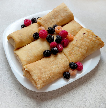 Pancakes with berries with filling on a white plate close-up on a white table with glass, top view, pancakes rolled up in an envelope, homemade foodの写真素材
