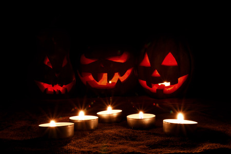 Halloween pumpkin blurred three jack-o'-lantern in the darkness on sackcloth among candles, focus on candlesの写真素材