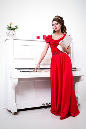Elegant woman in a long red dress is standing at the piano with unidentified notes in the handsの写真素材