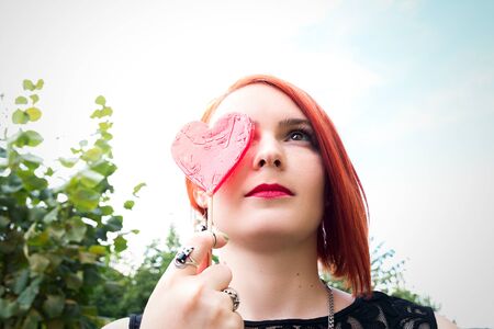 Beautiful red-haired girl covers her eyes with candy in the shape of heart