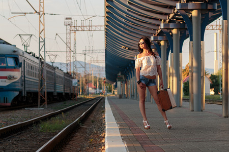 Lonely young brunette woman in sexy shorts with old suitcase on station platform with sunlight in...