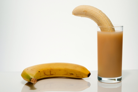 Ripe banana without peel from the tropics in a glass with banana juice and yellow ripe banana close by, on a light gradient background highlighted with space for textの写真素材