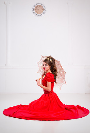 Beautiful elegant girl in a long red dress and shoes sits on the floor with an old vintage umbrella in a white roomの写真素材