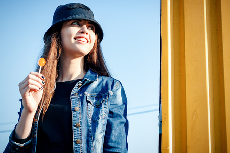Cheerful stylish young girl in hat, and jeans jacket with candy on stick, Chupa Chups, on sunny day standing on the street against tree with large portrait. With space for textの写真素材