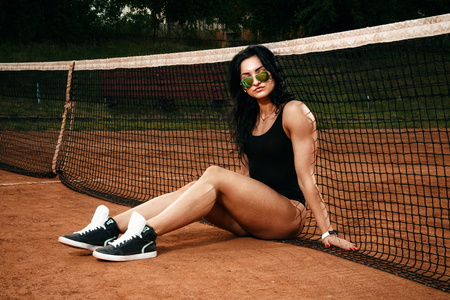 Beautiful young free sports sexy girl dressed in bodysuit, sneakers, with a smart ass and long legs, on the sports ground sits at the net on the tennis courtの写真素材
