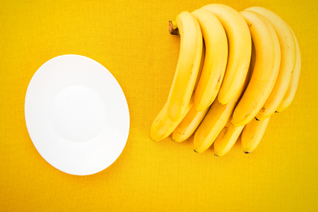 Bunch of yellow ripe bananas from the tropics with a white plate on a yellow fabric background with space for textの写真素材
