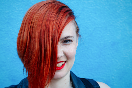 Bright portrait of a red-haired girl with freckles standing against a blue wall, female serious in a leather jacketの写真素材