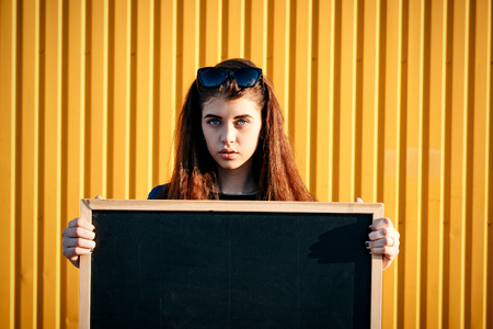 Cheerful stylish young girl in denim jacket, wearing  sunglasses, with chalkboard against yellow wall on sunny day. With space for textの写真素材
