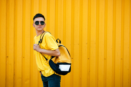 Stylish serious guy in a dirty yellow T-shirt and sunglasses and ripped jeans with a backpackの写真素材