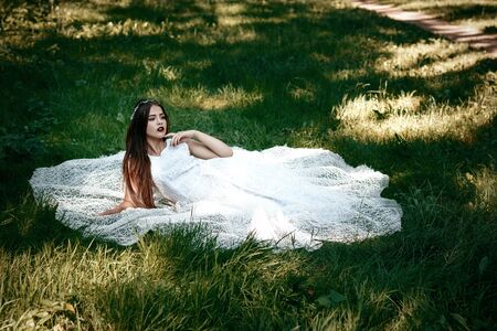 Woman or girl, a bride in a white wedding dress, sitting on the grass in the middle of a forest or park, happy photoの写真素材