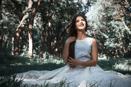 Woman or girl, a bride in a white wedding dress, sitting on the grass in the middle of a forest or park, happy photoの写真素材