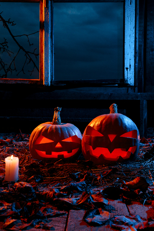 The concept of Halloween. Two luminous evil scary pumpkins, jack-lantern, with candles, leaves and eerie branches outside the window with a warm and cold blue light on the wooden floorの写真素材