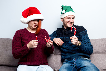Spirit of Christmas and new year. Concept of a holiday and days off. A loving couple in Santa Claus hats. The girl is sad with a broken candy, the guy is pleased with the coffee-grinder stickの写真素材