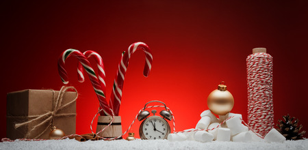 Christmas composition. Greeting card for new year alarm clock toys balls candy stick cinnamon marshmelow thread gift on snow on red background with place for greeting text の写真素材