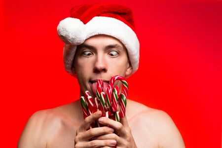Christmas new year concept food and sweets. Man in santa claus hat holding many candies stick. Stylish happy guy lick a lot of candy cone on red backgroundの写真素材