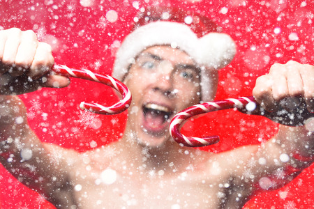 Christmas new year concept food and sweets. Man in santa claus hat holding two candies stick. Stylish happy guy smile with candy cone on red background. Focus on hook with snowflakesの写真素材