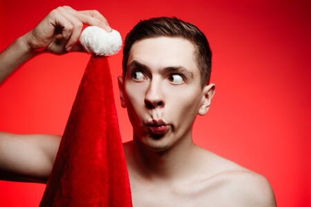 Christmas new year concept. Man with santa claus hat looking funny. foolish Guy with stylish hair on red background with dark brown eyesの写真素材