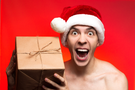 Christmas holiday presents. Crazy Man hold New year gift. Stylish guy in santa claus hat very happy on red background with torsoの写真素材