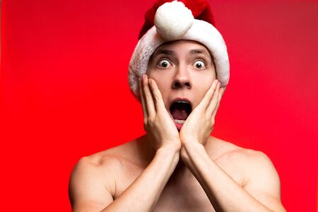Christmas new year concept. Man with santa claus hat looking scared. Stylish Guy looks like home alone on red backgroundの写真素材