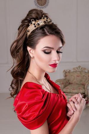 Elegant woman in long red dress standing in white room. Copy space. European tall girl with makeup. Beauty lifestyle concept.の写真素材