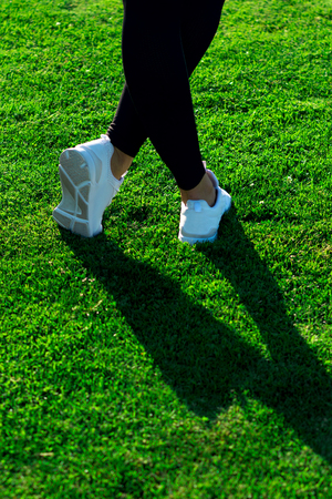 Sports Legs of young athletic woman in dark tights and light sneakers stay or walking along path among dense green grass in training. Copy space. Healthy lifestyle concept. European femaleの写真素材