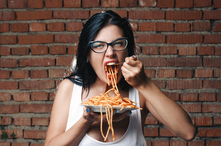 Sexy seductive girl in white T-shirt with plate of pasta and ketchup, with fork eats pasta. Erotic sucking noodles, standing against brick wall. Brunette in glasses love food and very hungryの写真素材