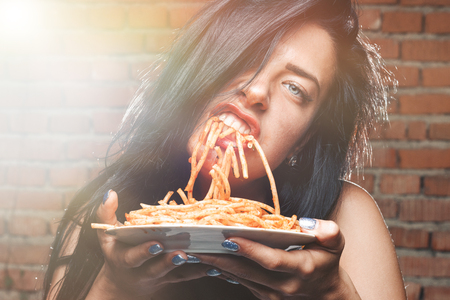 Sexy seductive girl in white T-shirt with plate of pasta and ketchup, without fork eats pasta. Erotic sucking noodles, standing against brick wall. Brunette with blue eyes love food and very hungryの写真素材