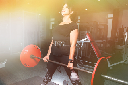 Woman plus size in gym doing exercises with barbell powerlift, female XXL losing weight, fat model working with her body in fitness hall, healthy lifestyle and dietの写真素材