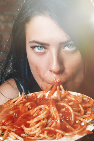 Sexy seductive girl in white T-shirt with plate of pasta and ketchup, without fork eats pasta. Erotic sucking noodles, standing against brick wall. Brunette with blue eyes love food and very hungryの写真素材
